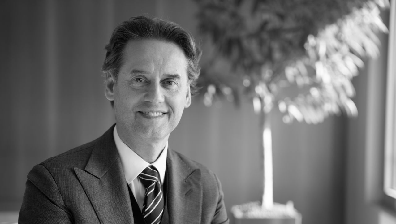 Un uomo sorridente in abito formale e cravatta a righe siede in una stanza ben illuminata con una pianta visibile sullo sfondo. L'immagine è in bianco e nero.