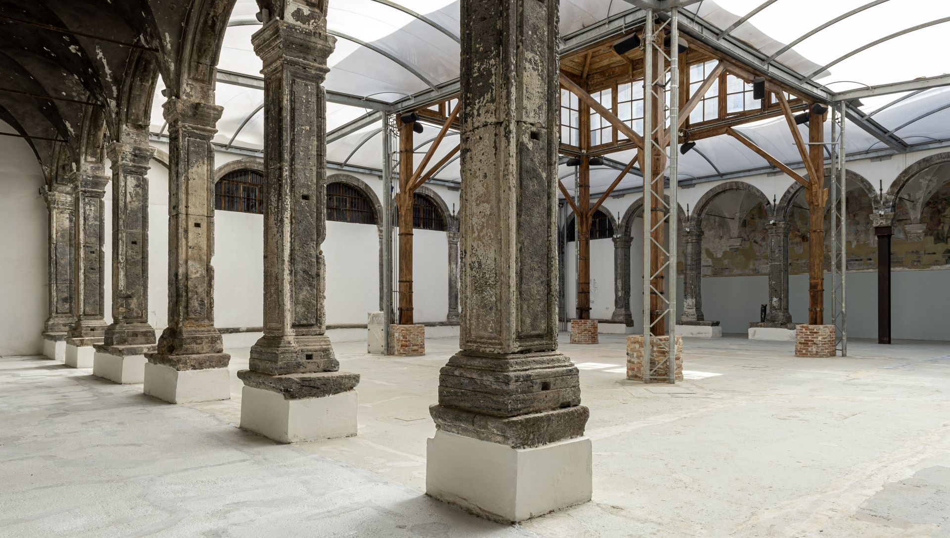 Un interno architettonico spazioso e storico con alte colonne e archi in pietra sotto un soffitto curvo e traslucido. Il pavimento è di colore chiaro e spoglio, e alcune travi sono rinforzate con basi in mattoni. Le pareti mostrano segni di età e di parziale restauro.