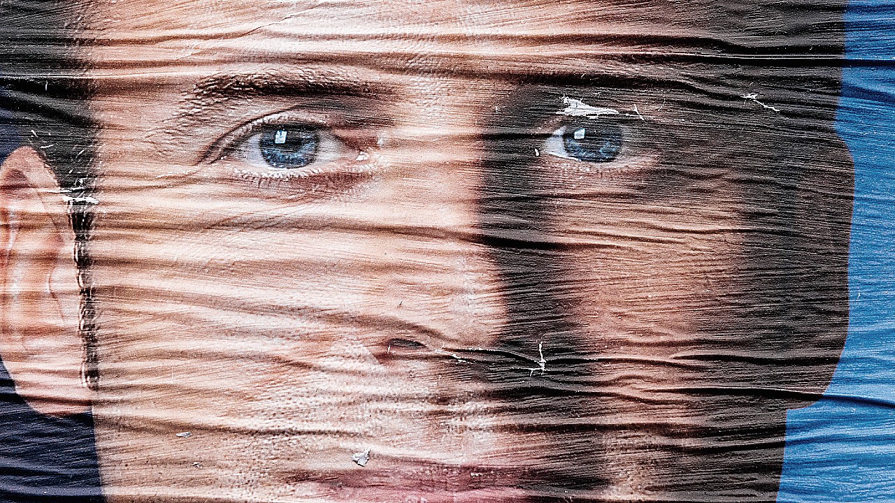 Un primo piano del volto di un uomo stampato su un poster sgualcito e strappato, incentrato su occhi e naso. Il poster è rovinato, con linee orizzontali e pieghe che ne distorcono i lineamenti.