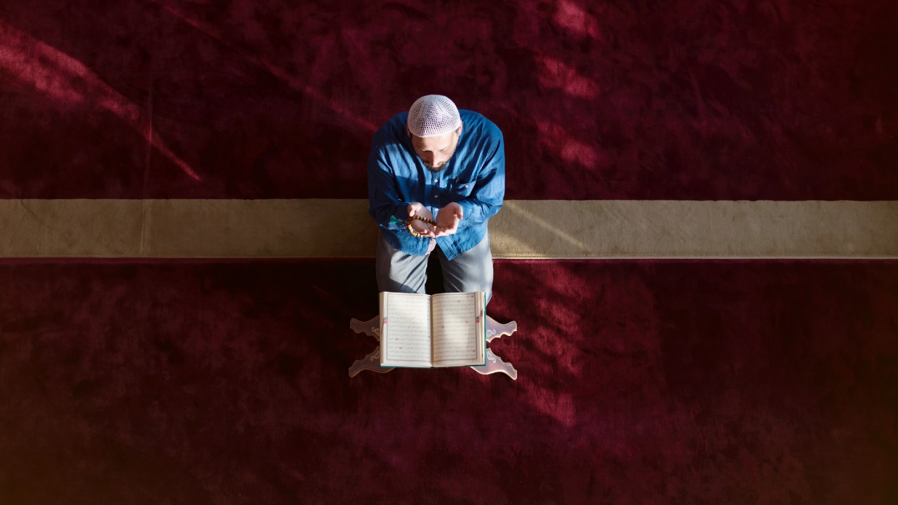 Una persona che indossa una camicia blu e un berretto bianco è inginocchiata su un tappeto rosso, tenendo le mani giunte in preghiera. Un libro aperto è appoggiato su un supporto di fronte a loro. La scena è illuminata da una luce naturale e soffusa dall'alto.