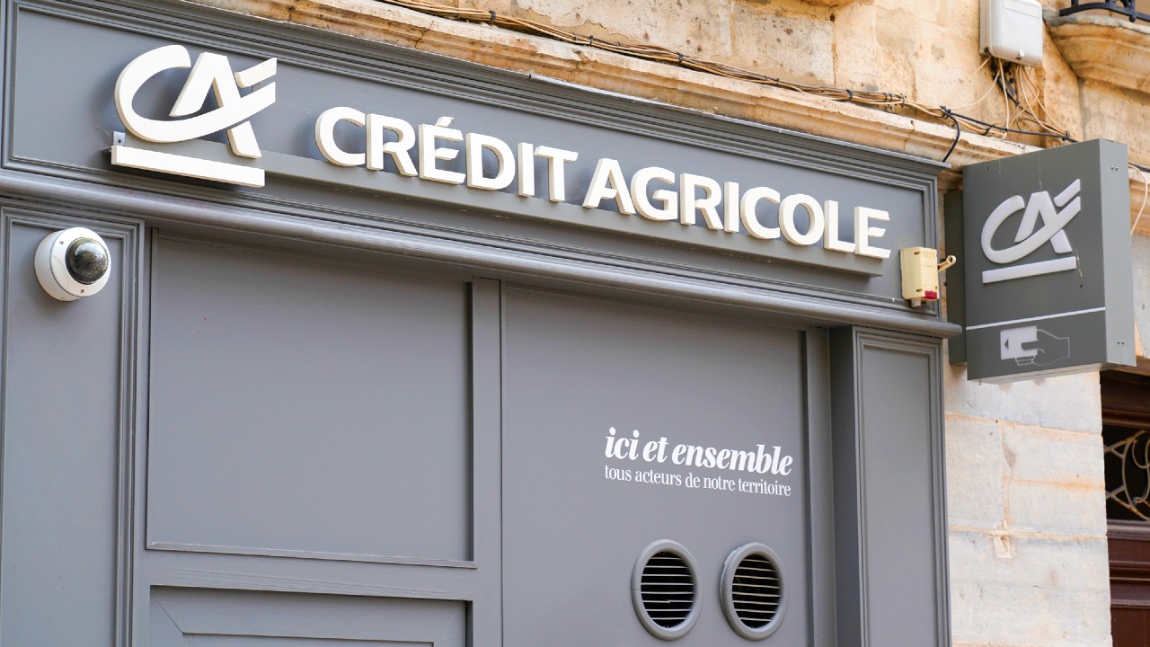 Facciata di una filiale del Crédit Agricole con segnaletica grigia e bianca. Le parole "ici et ensemble, vos acteurs de notre territoire" sono visualizzate in francese. Le telecamere di sicurezza sono visibili sul muro esterno.
