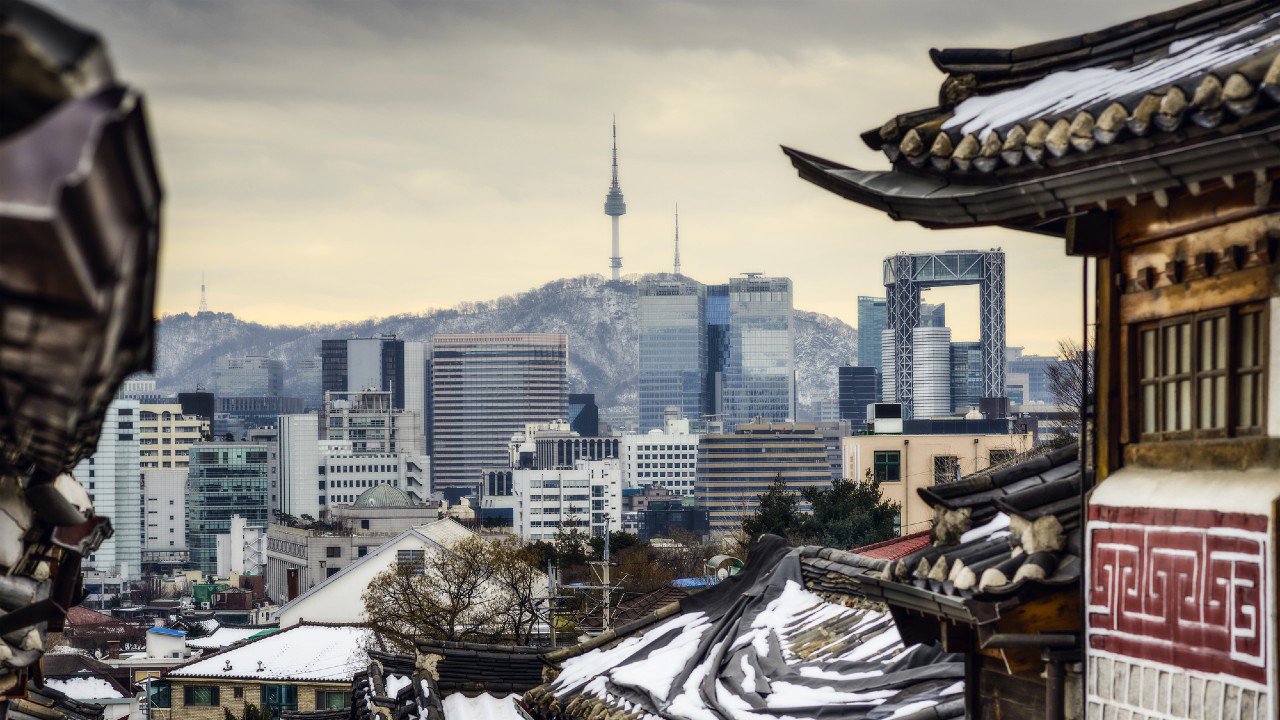 Un paesaggio urbano di Seul, in Corea del Sud, presenta i tradizionali tetti degli hanok in primo piano e moderni grattacieli sullo sfondo, con la Namsan Seoul Tower visibile in cima a una collina sotto un cielo nuvoloso.