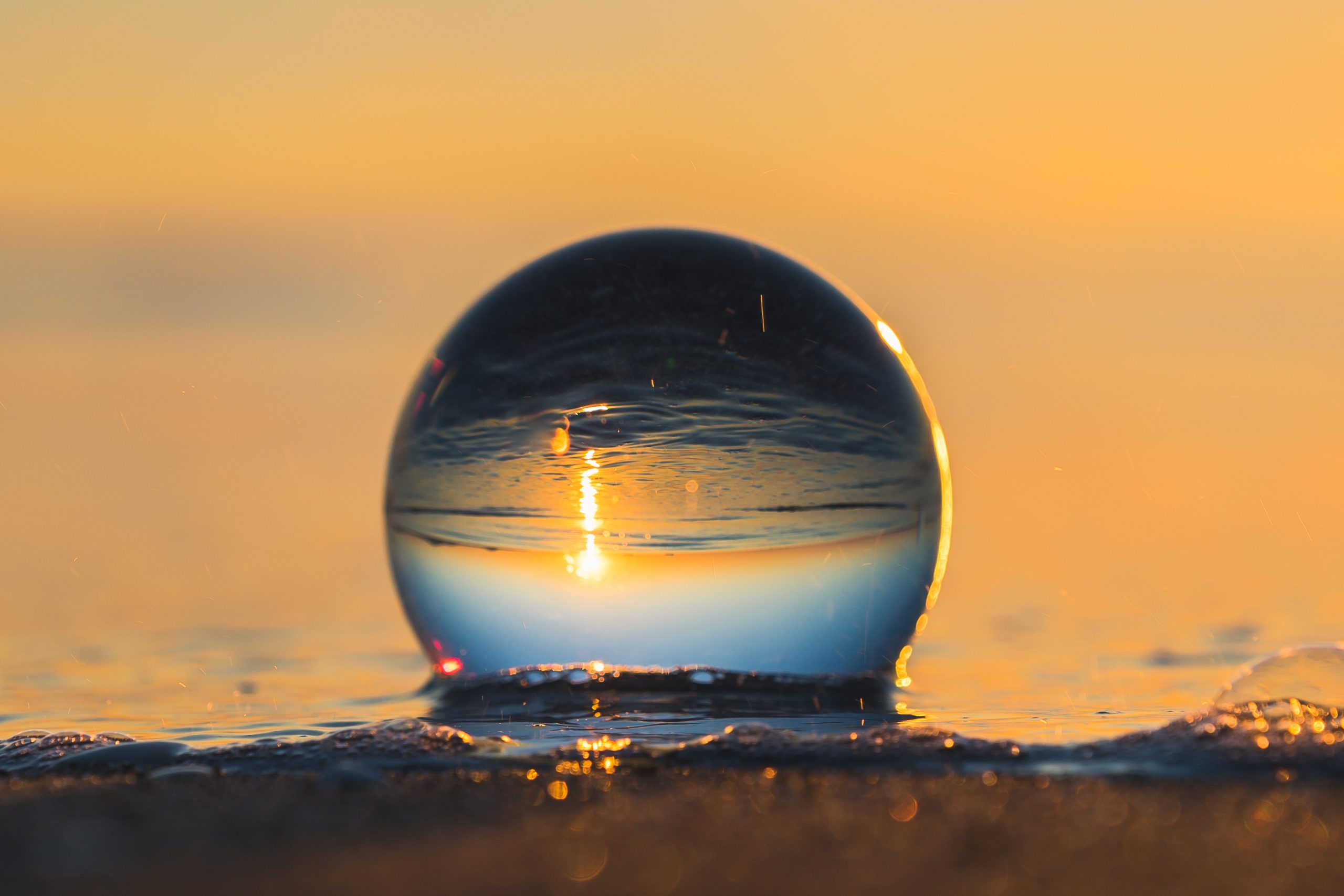 Una sfera di cristallo posta sulla sabbia riflette un tramonto sull'oceano, proprio come una maturità target che raggiunge la sua sorprendente conclusione. Il cielo si illumina di sfumature di arancione e giallo, con il sole parzialmente visibile attraverso la sfera trasparente, creando un effetto specchiato dell'orizzonte e delle onde.