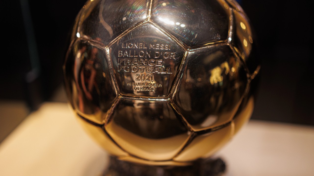 Primo piano di un trofeo dorato del Pallone d'Oro con inciso il testo "Lionel Messi, Pallone d'Oro, France Football, 2021". Il trofeo ha la forma di un pallone da calcio ed è posizionato su un supporto.
