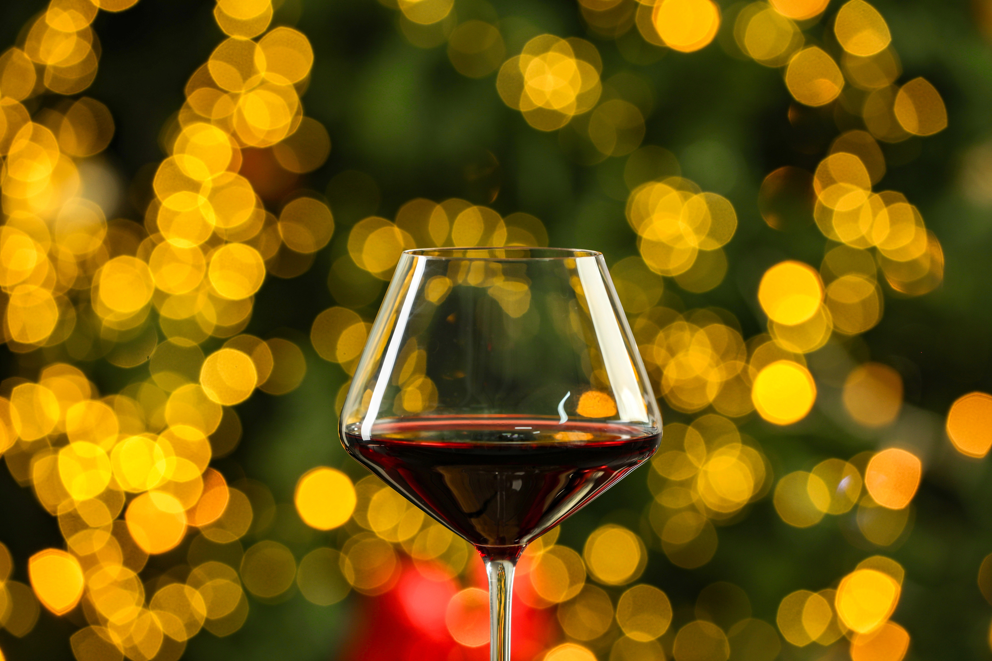 Un bicchiere di vino rosso è posto al centro di uno sfondo di luci bokeh gialle calde e luminose, creando un'atmosfera festosa ed elegante.