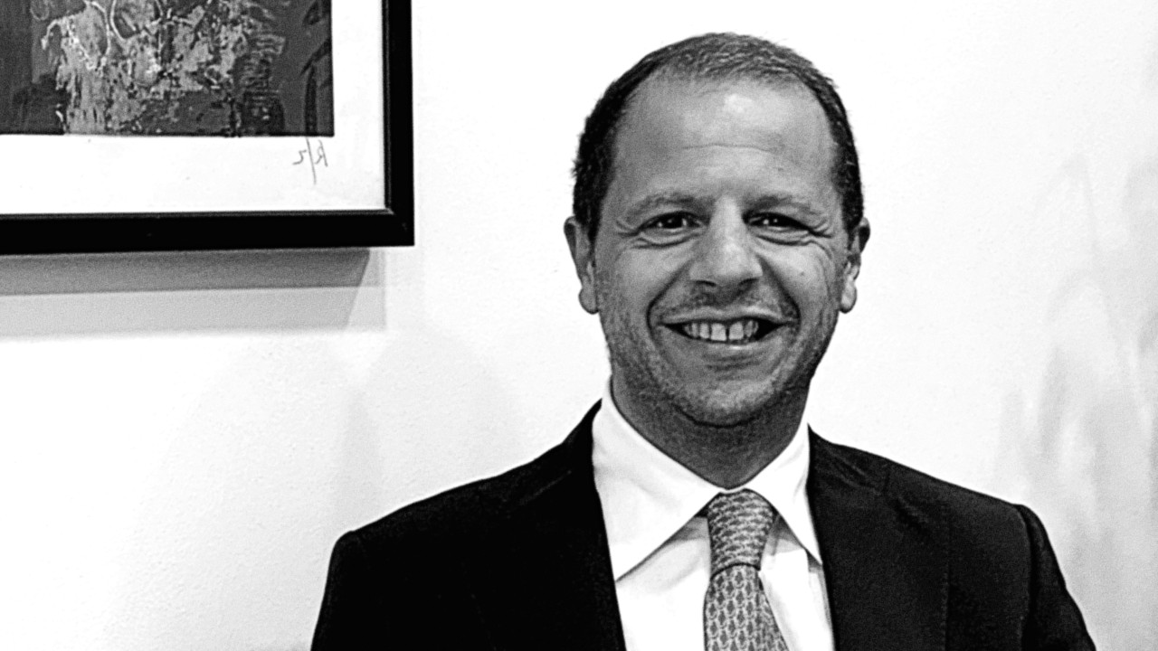 Un uomo in giacca e cravatta sorride alla telecamera in una foto in bianco e nero. C'è un'opera d'arte incorniciata parzialmente visibile sul muro sullo sfondo.
