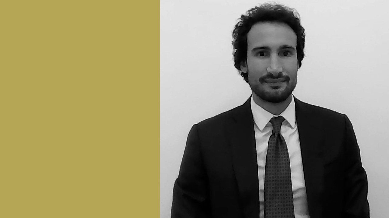 Immagine in bianco e nero di un uomo in giacca e cravatta, Tommaso Marchetti, seduto su uno sfondo bianco semplice. I suoi corti capelli ricci e la barba ben curata evidenziano un aspetto professionale. C'è un rettangolo dorato sul lato sinistro dell'immagine.