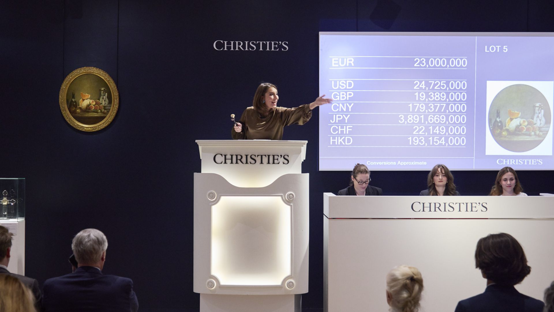 Un banditore è in piedi su un podio con la scritta "Christie's", mentre conduce un'asta. Uno schermo mostra varie valute e importi. I partecipanti sono seduti di fronte al palco, dove altri due sono presenti a una scrivania. Un dipinto è visibile sul muro.