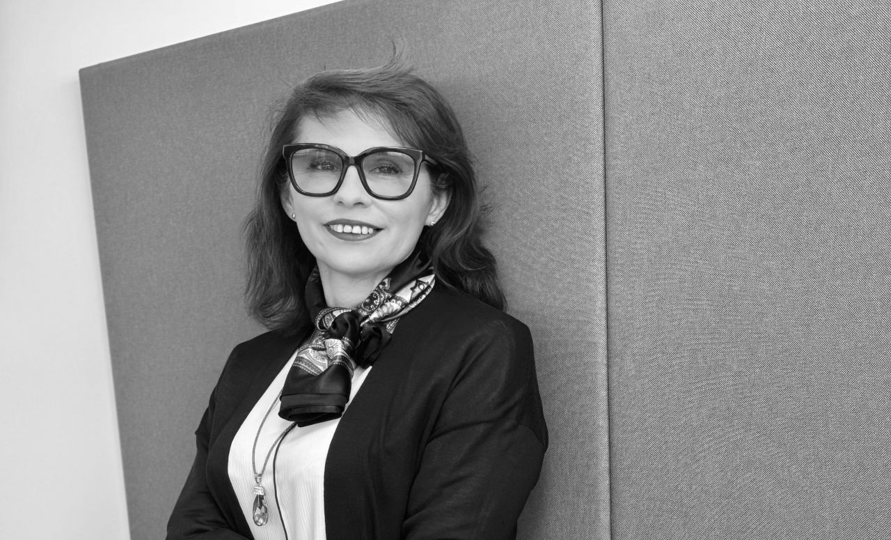 Una donna sorridente con grandi occhiali e capelli lunghi fino alle spalle è appoggiata a una parete ruvida. Indossa una giacca scura, una sciarpa fantasia e una collana con ciondolo. L'immagine è in bianco e nero.