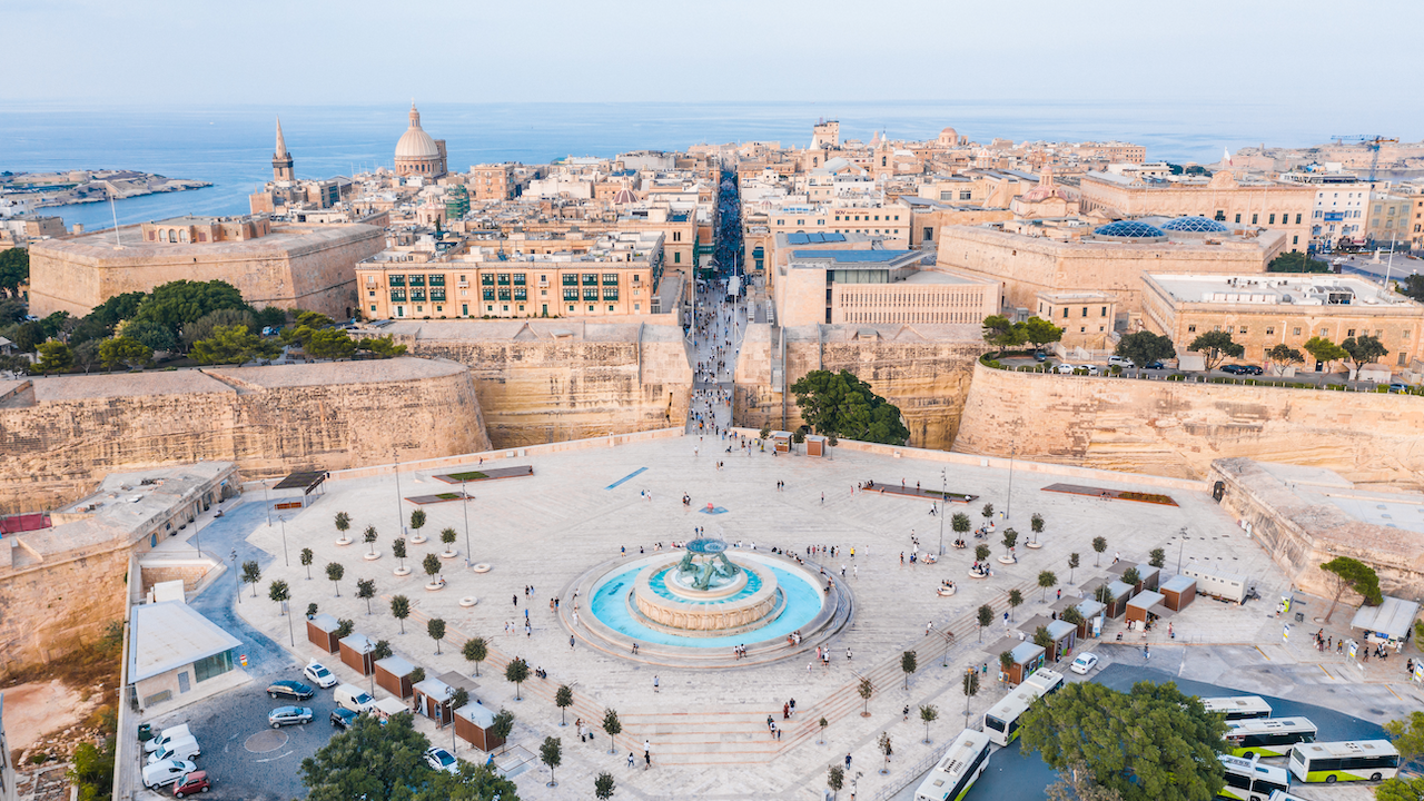 Vista aerea di La Valletta, Malta, che mostra la sua architettura storica e le strade trafficate. Una fontana prominente si trova in primo piano, circondata da persone e alberi, con il paesaggio urbano e la costa che si estendono sullo sfondo.