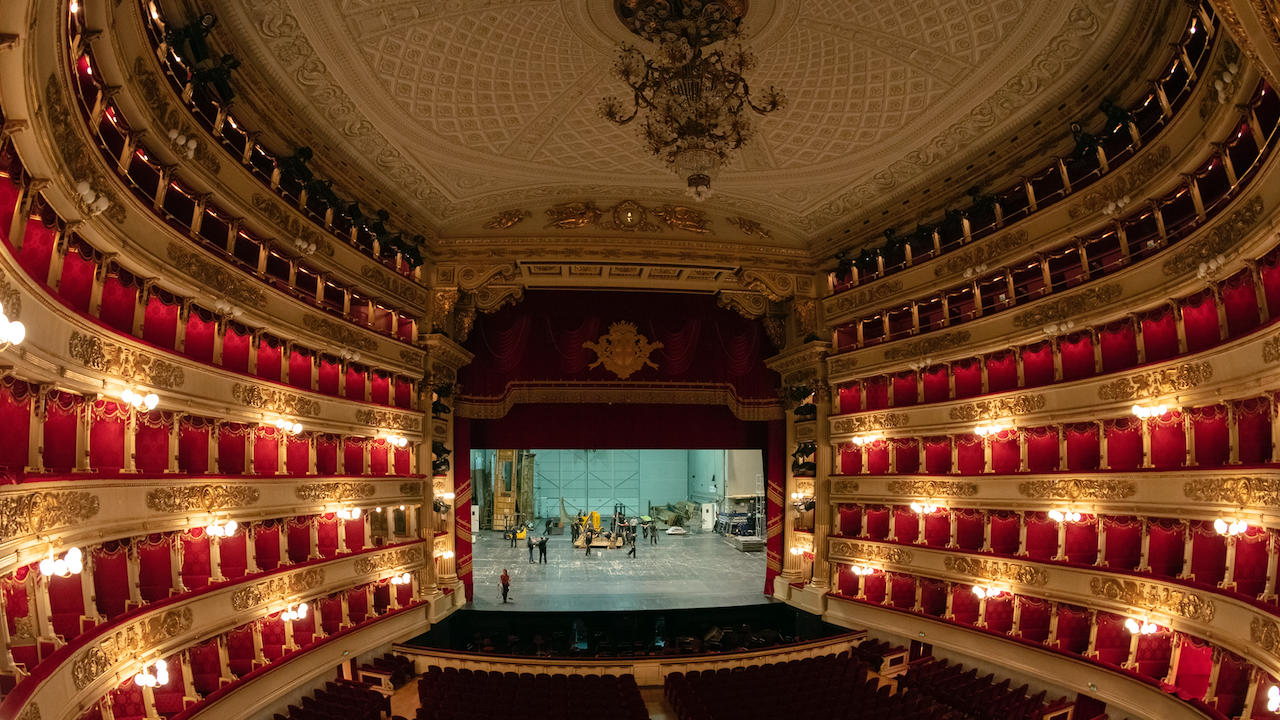 Interno di un teatro ornato con sedili in velluto rosso, dettagli dorati e un grande lampadario. Il palco è allestito con persone che sistemano oggetti di scena, creando un sorprendente contrasto con l'ambiente opulento. Balconi si innalzano su entrambi i lati con un'illuminazione spettacolare.