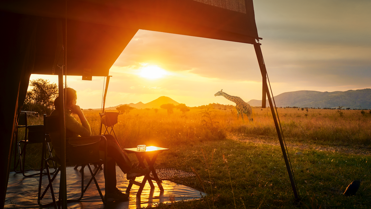 Una persona è seduta sotto una tenda Turisanda, godendosi la vista del tramonto in un paesaggio di savana. Un tavolo con un drink è appoggiato lì vicino, mentre una giraffa è in piedi in lontananza, stagliata contro il caldo bagliore del sole al tramonto.