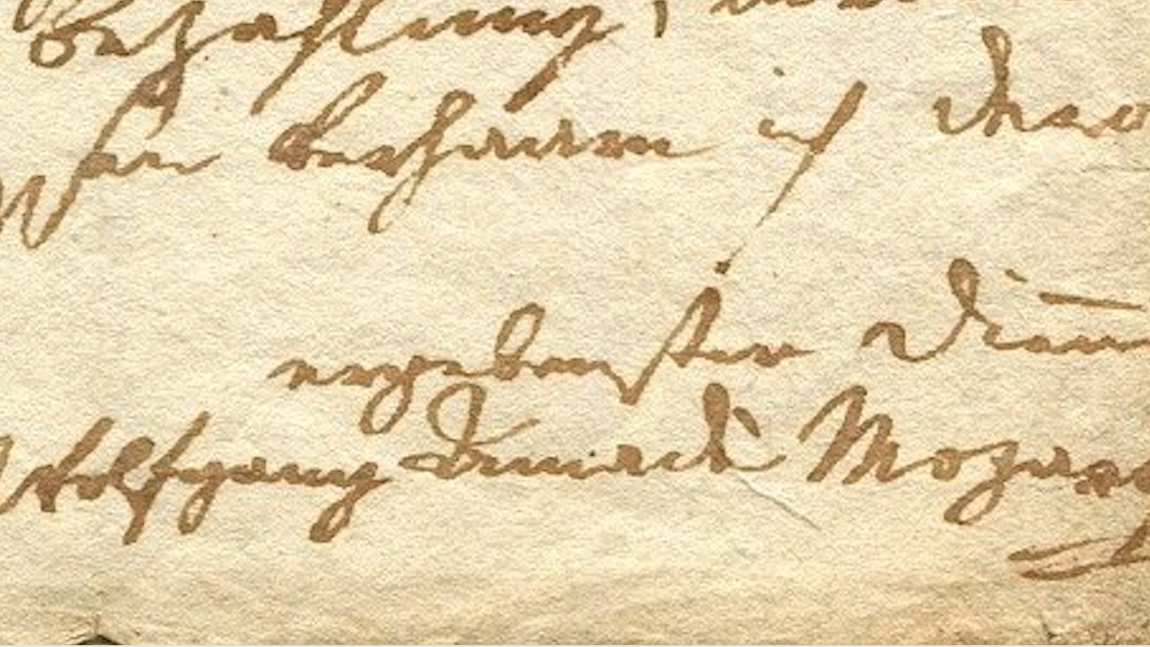 Primo piano di un documento manoscritto antico con scrittura corsiva in inchiostro marrone su carta beige. Il testo include il nome "Wolfgang Amadeus Mozart". La scrittura è ornata e parzialmente sbiadita, con texture della carta e pieghe visibili.