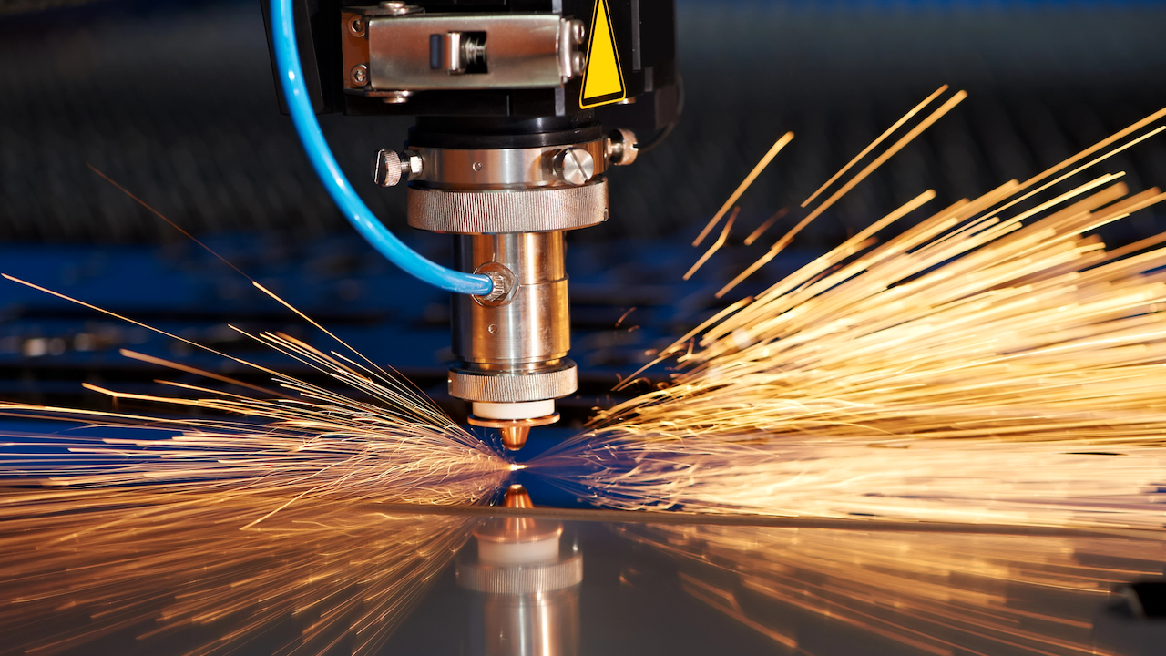 Primo piano di una macchina per il taglio laser CNC in azione. La macchina emette scintille luminose mentre taglia con precisione una lamiera. Un tubo flessibile blu e componenti metallici sono visibili nell'impostazione, evidenziando la produzione industriale.