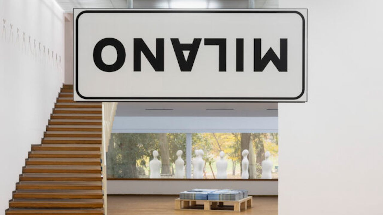 L'interno di una galleria d'arte moderna con un grande cartello capovolto che recita "MILANO". Sulla sinistra c'è una scala di legno e sullo sfondo si vedono figure simili a manichini attraverso una finestra. Una collezione di libri è esposta su un pallet.