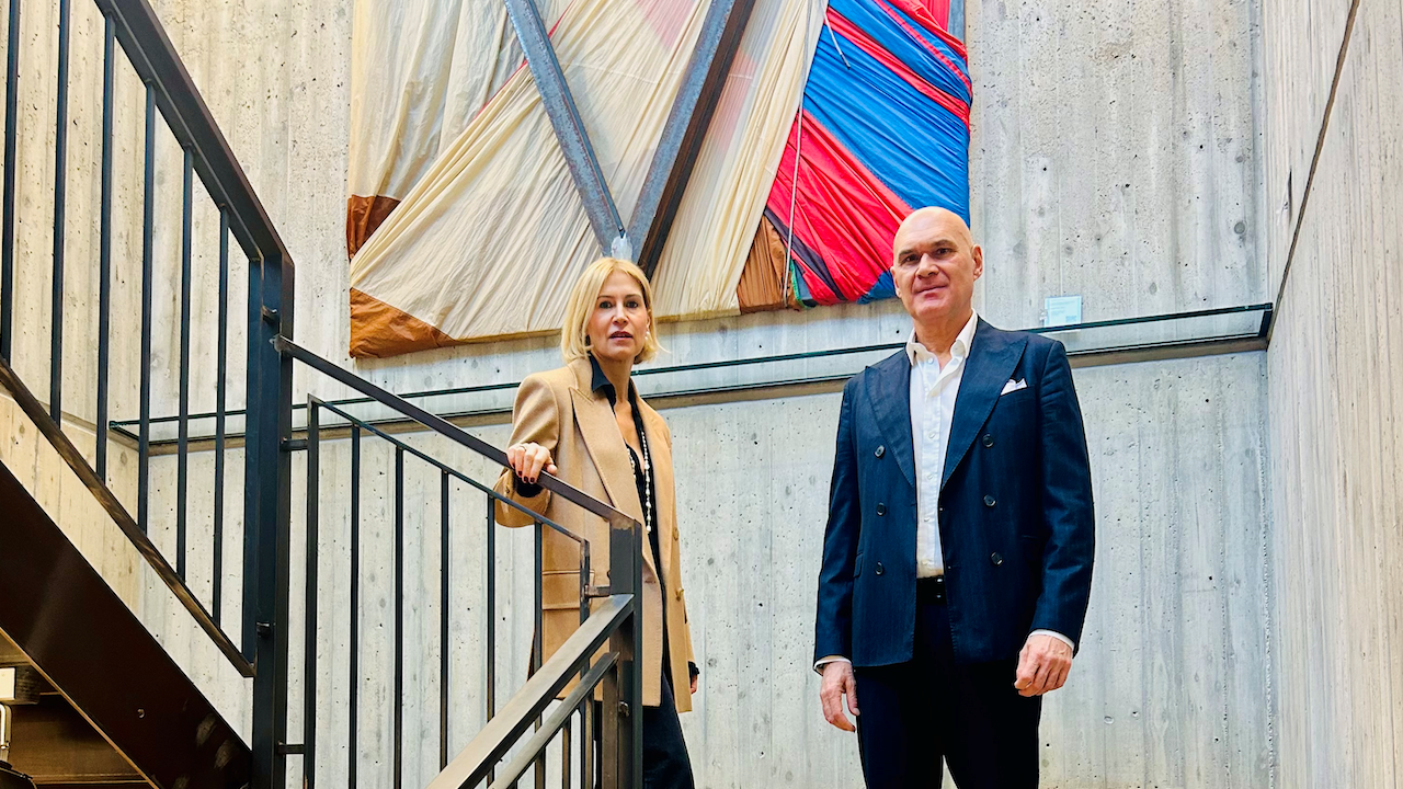 Una donna e un uomo sono in piedi su una scalinata di fronte a un muro di cemento decorato con arte astratta in tessuto. La donna indossa un cappotto beige e pantaloni neri, mentre l'uomo indossa un completo blu navy. I colori vivaci dell'arte contrastano con i toni neutri dello spazio.