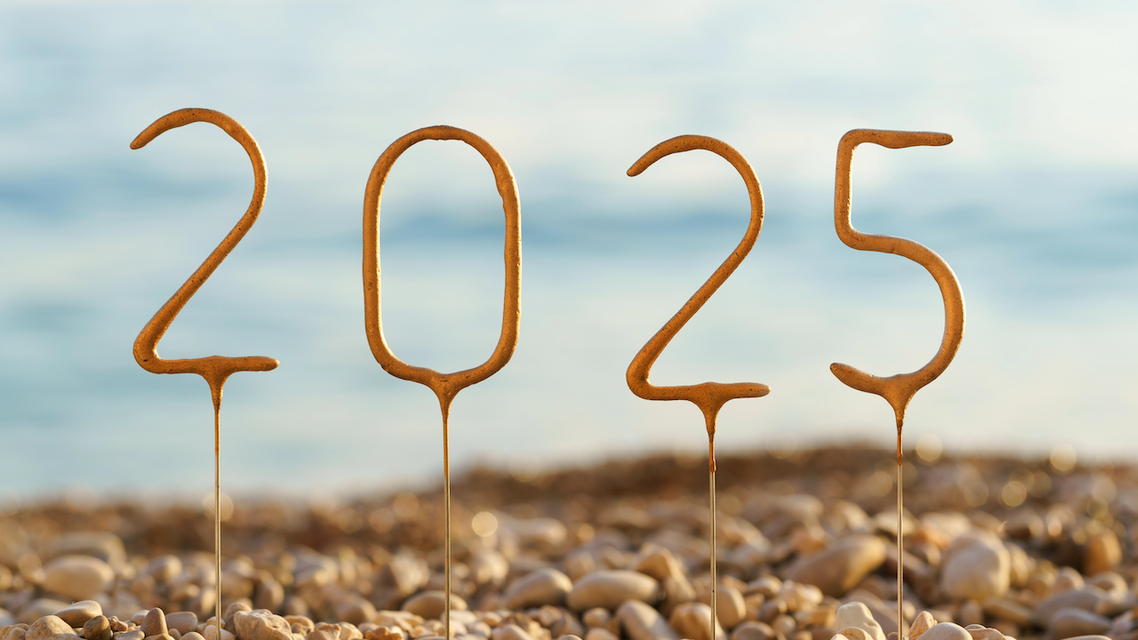 I numeri "2023" sono formati con la sabbia e sono posizionati verticalmente su una spiaggia, con ciottoli in primo piano e un mare e un cielo sfocati sullo sfondo.