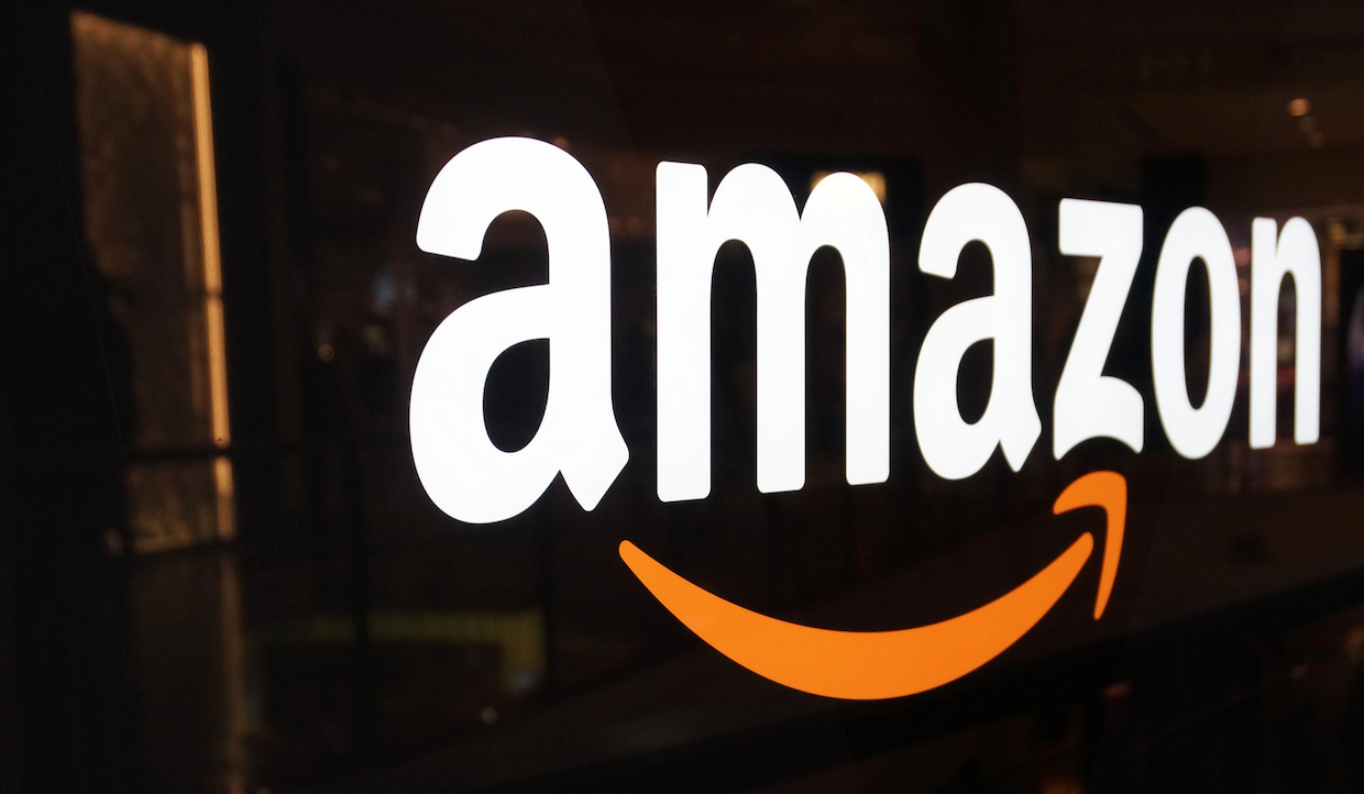 L'immagine mostra un primo piano del logo Amazon in lettere bianche con una freccia arancione sotto, che forma un sorriso. Lo sfondo è scuro, evidenziando l'aspetto luminoso del logo.