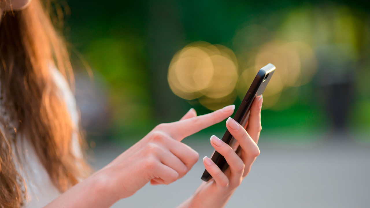 Primo piano di una persona che usa uno smartphone all'aperto, probabilmente mentre interagisce con il suo IT wallet. La persona ha i capelli lunghi e sta toccando lo schermo del telefono con l'indice. Lo sfondo è sfocato con tonalità verdi e gialle, suggerendo un ambiente naturale.