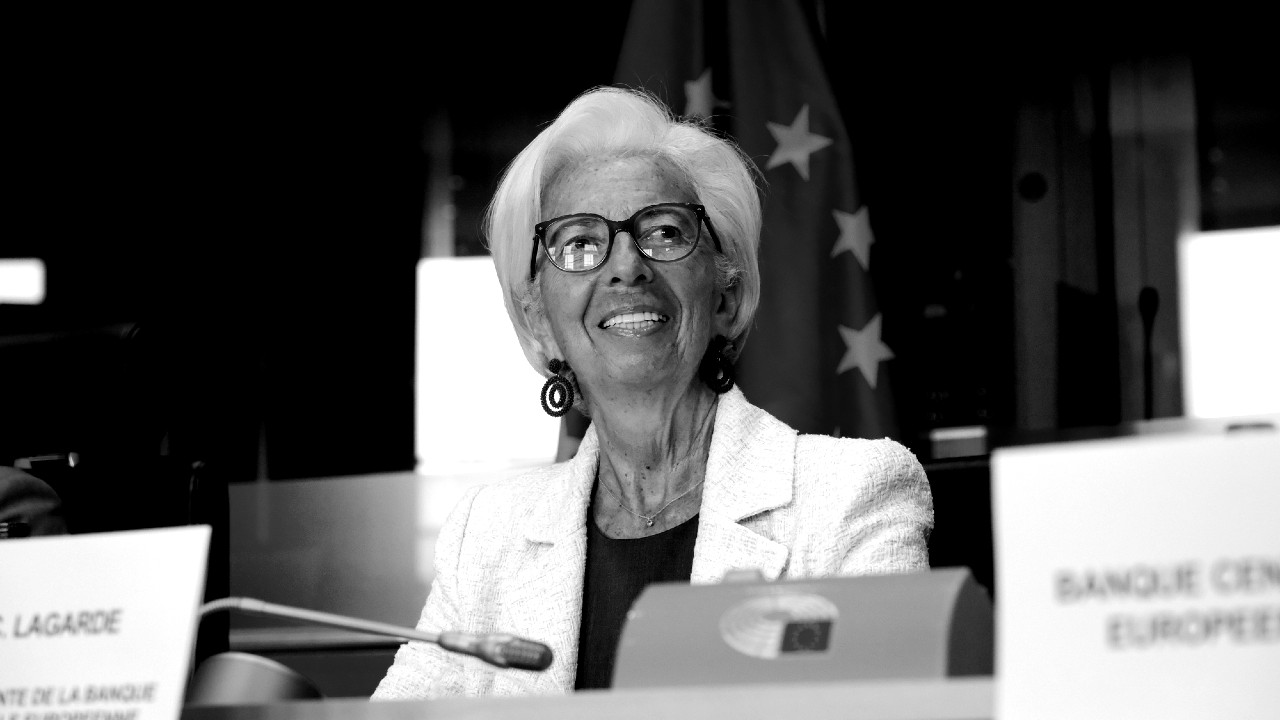 Una persona con i capelli bianchi corti e gli occhiali sta parlando a una conferenza. Indossa una giacca bianca e degli orecchini. Dietro di loro, è parzialmente visibile una bandiera dell'Unione Europea.