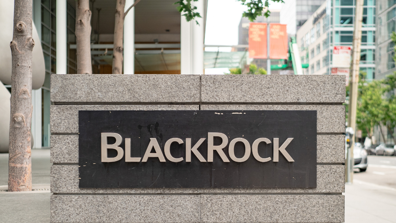Un grande cartello in pietra mostra con orgoglio l'iconico nome "BlackRock" di fronte a un elegante e moderno edificio per uffici. Sullo sfondo, un mix di rigogliosi alberi verdi e imponenti edifici cittadini completa la scena urbana.