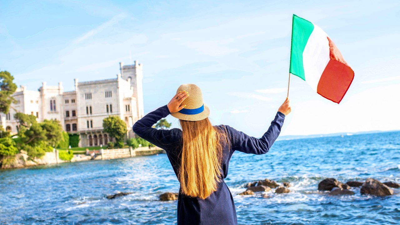 Una donna con i capelli lunghi, che indossa un cappello e un vestito blu, è in piedi vicino al mare con una bandiera italiana in mano. Sullo sfondo c'è uno storico castello bianco vicino all'acqua. Il cielo è limpido e azzurro, creando una pittoresca scena costiera.
