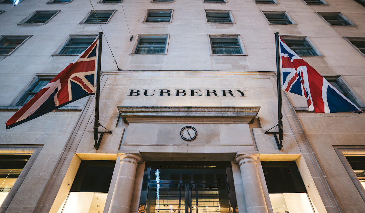 Una facciata di un edificio con il logo Burberry sopra un ingresso. Due bandiere Union Jack sono esposte su entrambi i lati dell'ingresso. C'è un piccolo orologio al centro sopra la porta.
