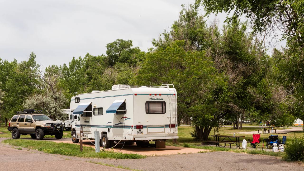 Un camper e un suv sono parcheggiati in uno dei tanti campeggi circondati da rigogliosi alberi verdi. Un tavolo da picnic con sedie rosse e blu è lì vicino e un'area erbosa si estende sullo sfondo sotto un cielo nuvoloso.