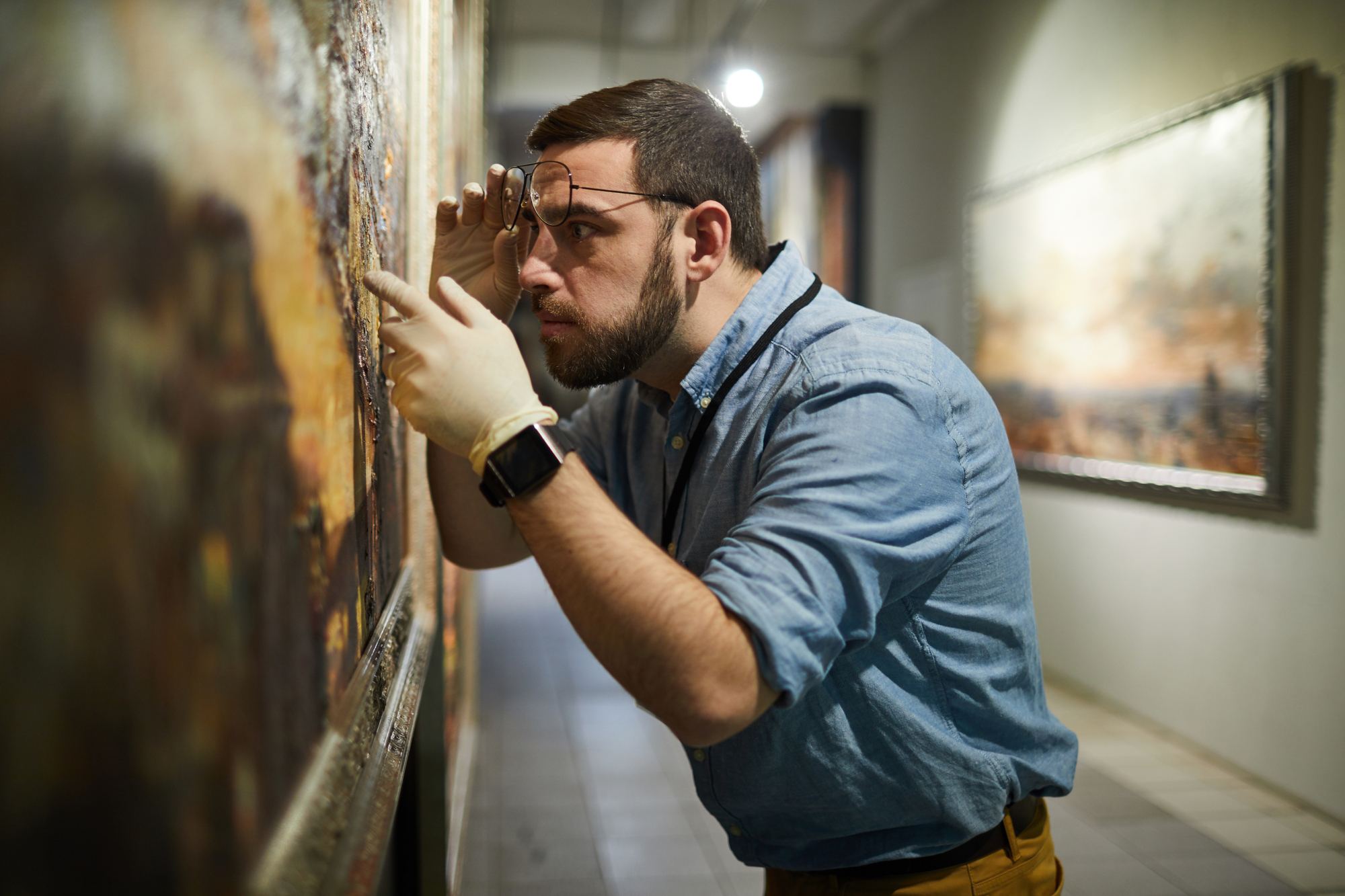 Un uomo con barba e occhiali, che indossa guanti e una camicia blu, esamina attentamente un dipinto in una galleria d'arte. Si sporge per ispezionare i dettagli, con altre opere d'arte visibili sullo sfondo.