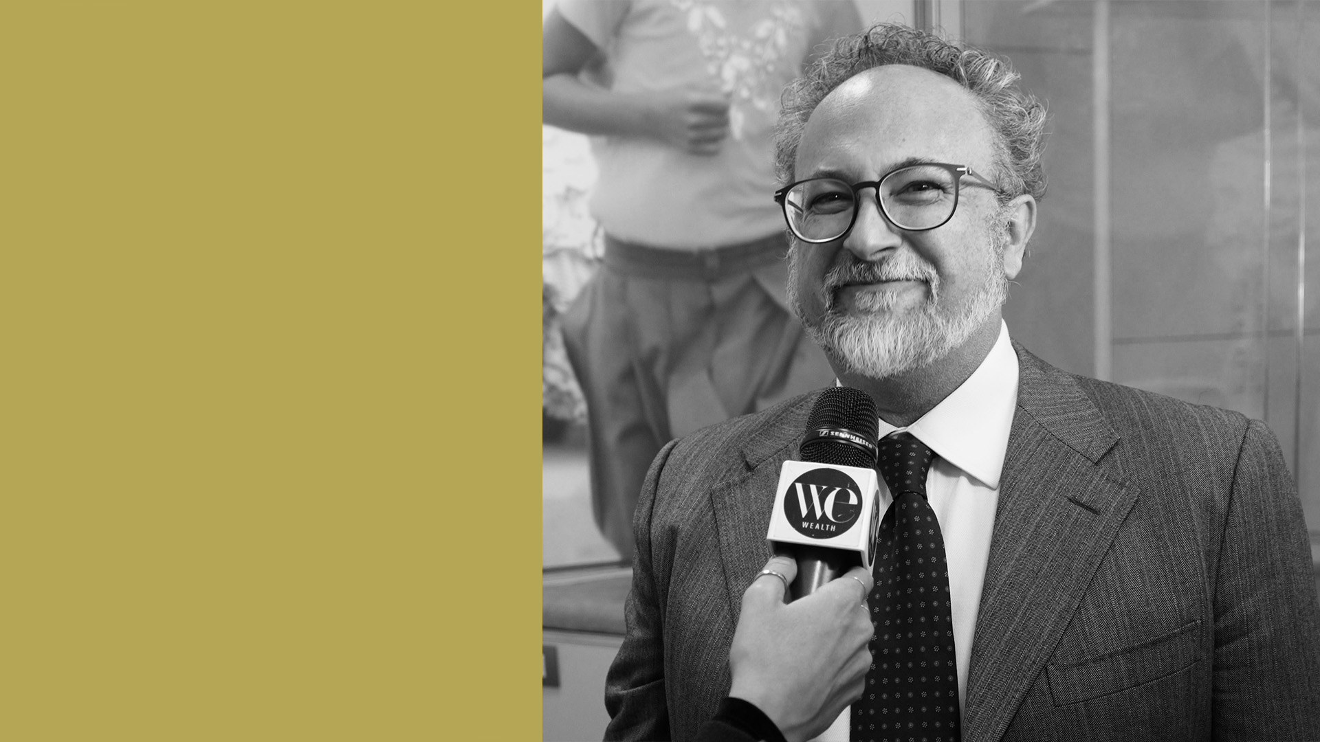 Una persona in giacca e cravatta sorride mentre viene intervistata da qualcuno che tiene in mano un microfono con la scritta "WC News". L'immagine è in bianco e nero, con una sezione giallo senape sul lato sinistro.