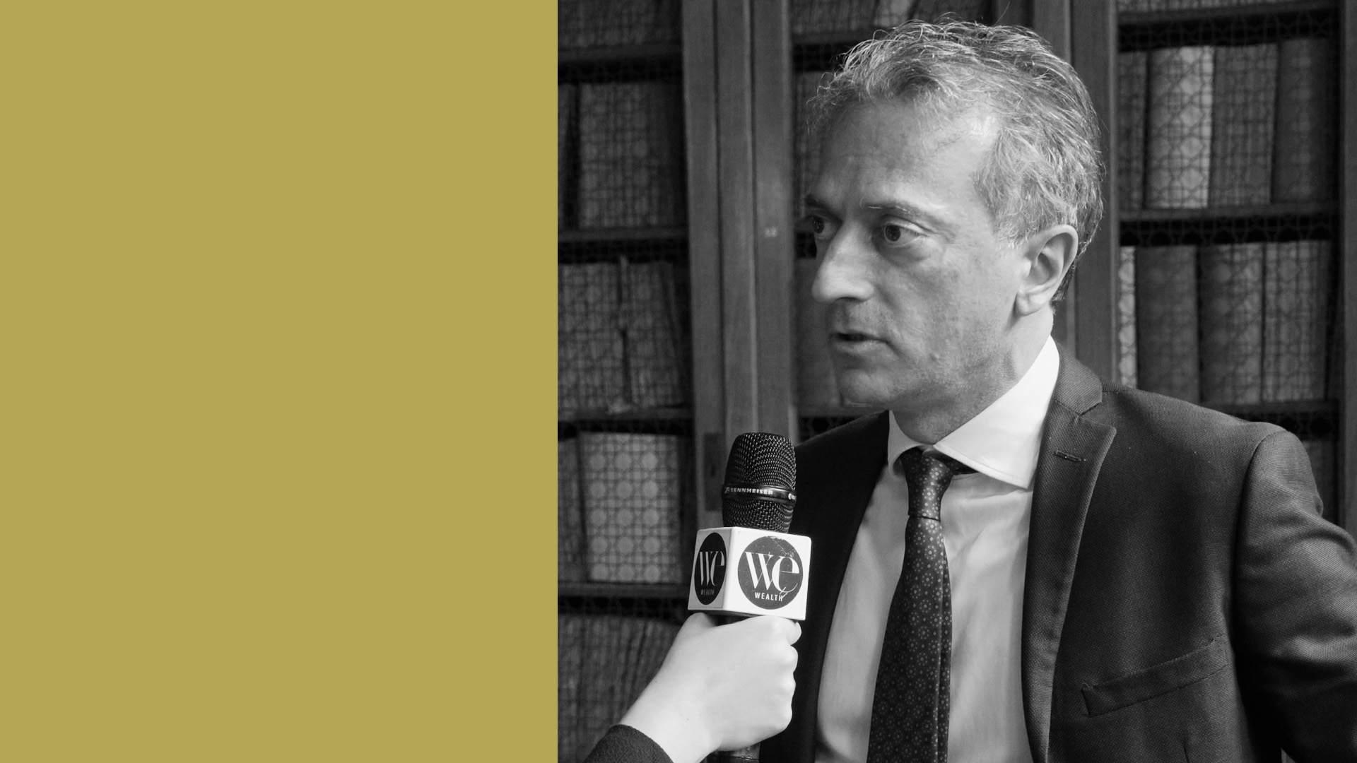 Andrea Testa di American Express, in giacca e cravatta viene intervistata in un ambiente chiuso. Una mano tiene un microfono con la scritta "noi" vicino al viso della persona, come se stesse parlando dei benefit dell'American Express. Lo sfondo mostra scaffali pieni di libri, mentre il lato sinistro dell'immagine presenta un solido colore giallo senape.