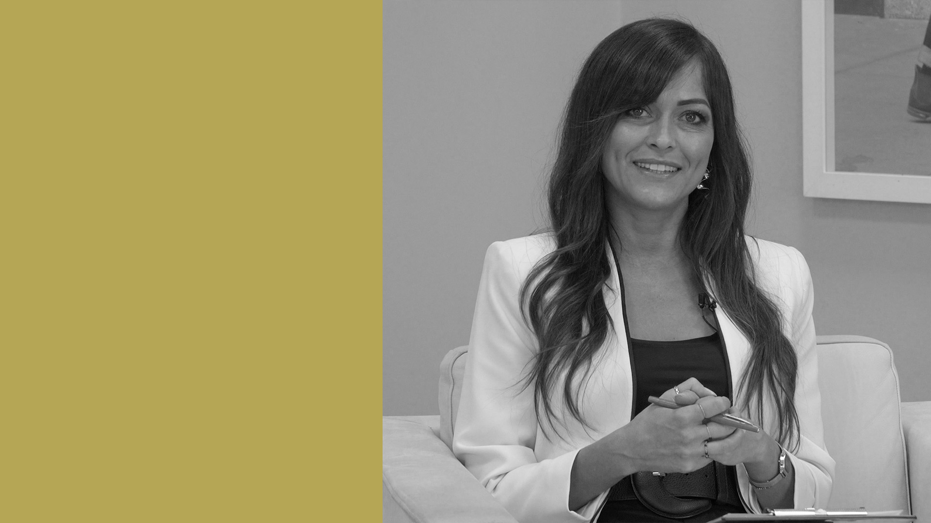 Una donna con lunghi capelli scuri in blazer bianco è seduta su un divano e sorride alla telecamera. Si chiama Chiara Samorì, giornalista di We Wealth. Il lato sinistro dell'immagine ha uno sfondo giallo senape uniforme. Sembra rilassata e sicura di sé.