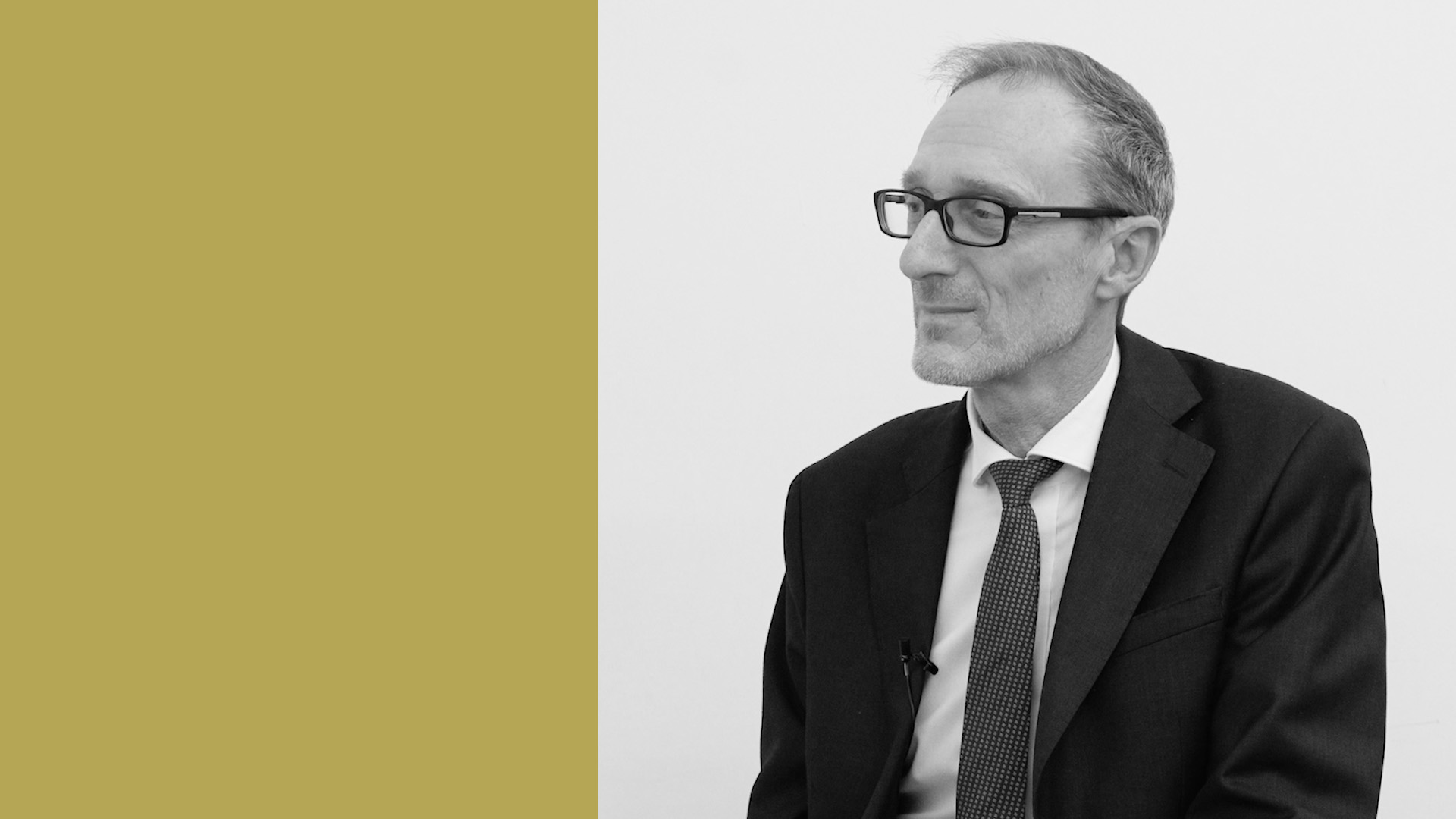 Un uomo in giacca e occhiali siede su uno sfondo bianco, il suo nome è Gianbattista Geroldi, ad per l'Italia di Objectway. Guarda pensieroso di lato. Accanto, un pannello color senape occupa il terzo sinistro dell'immagine, aggiungendo un contrasto vibrante a questo ritratto moderno.