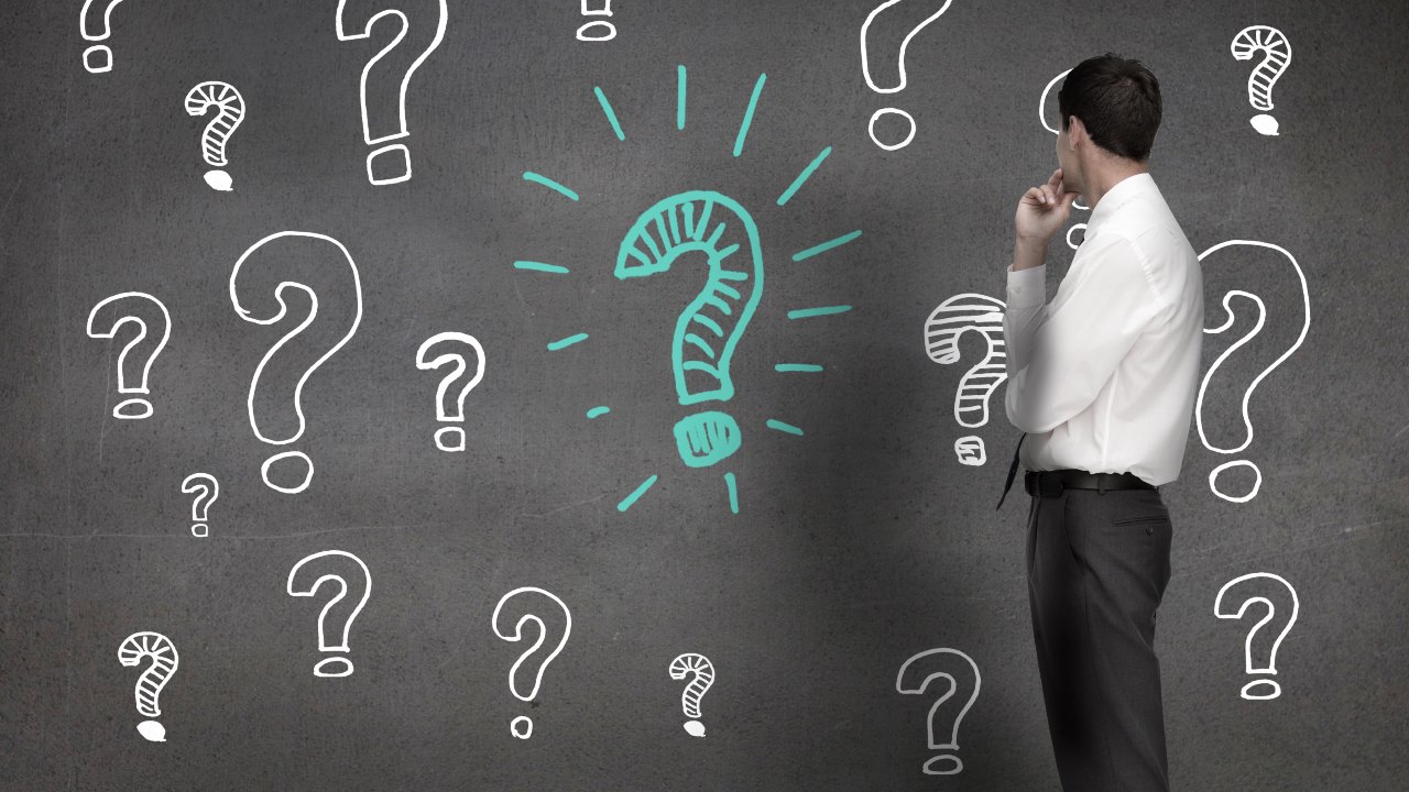 Un uomo con una camicia bianca e pantaloni grigi è in piedi, pensieroso, con una mano sul mento, di fronte a un muro grigio decorato con numerosi punti interrogativi bianchi e un prominente punto interrogativo blu-verde, e riflette sulle complessità delle tipologia di società da costiuire.