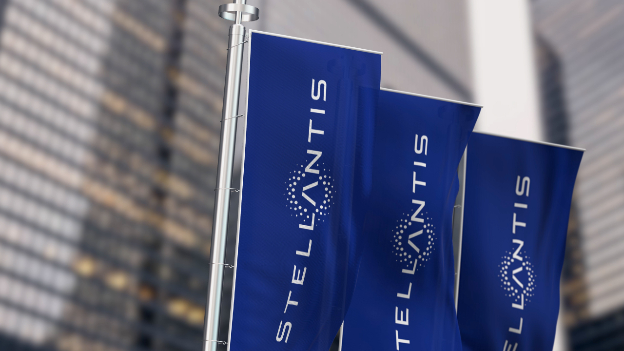 Tre bandiere blu con il logo e il nome Stellantis sono esposte su pennoni su uno sfondo di moderni edifici in vetro. Il logo include un motivo circolare a punti che circonda la lettera "A".