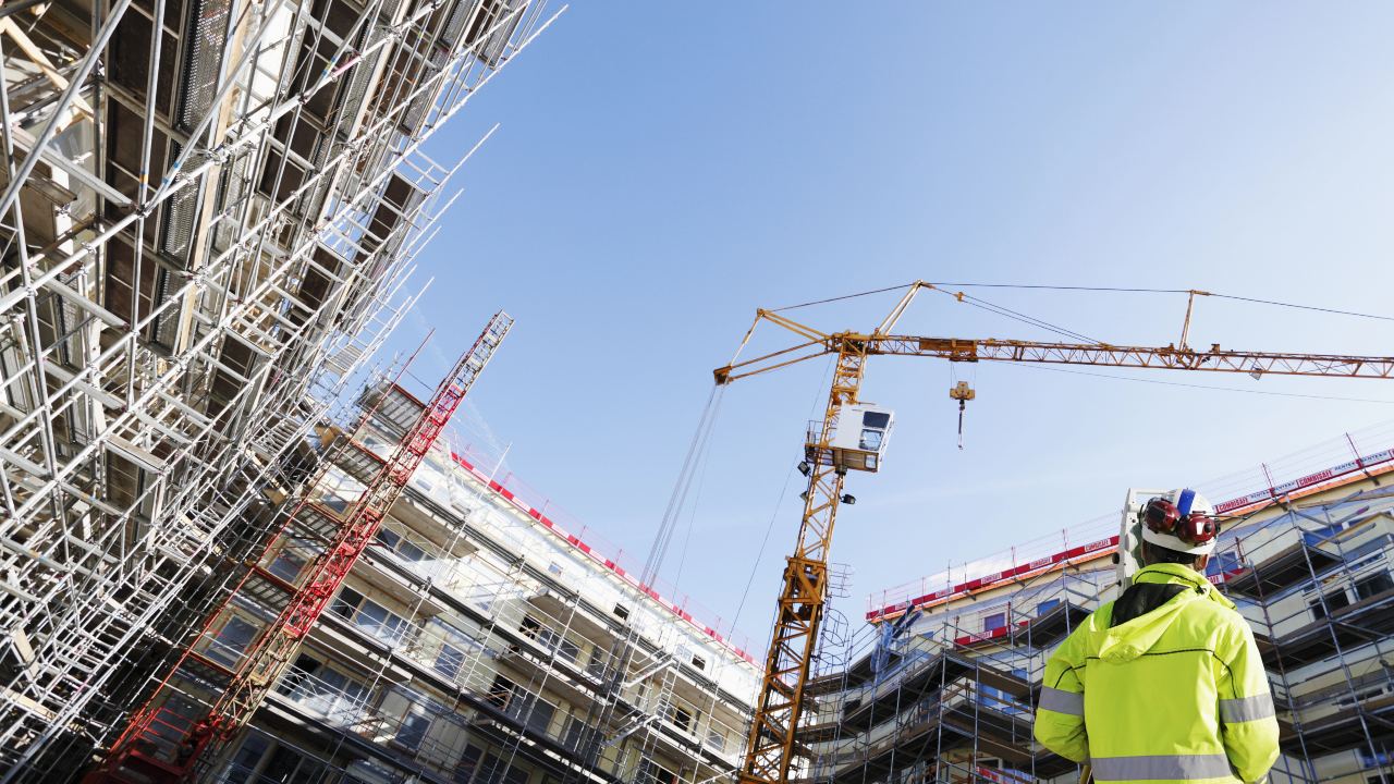 Immagine di un cantiere con impalcature che circondano gli edifici e una gru che si staglia contro il cielo azzurro e terso. Sulla destra, un operaio con una giacca gialla brillante e un casco è immobile e riprende l’idea dell’edilizia ferma in attesa del Ddl Salva Milano.