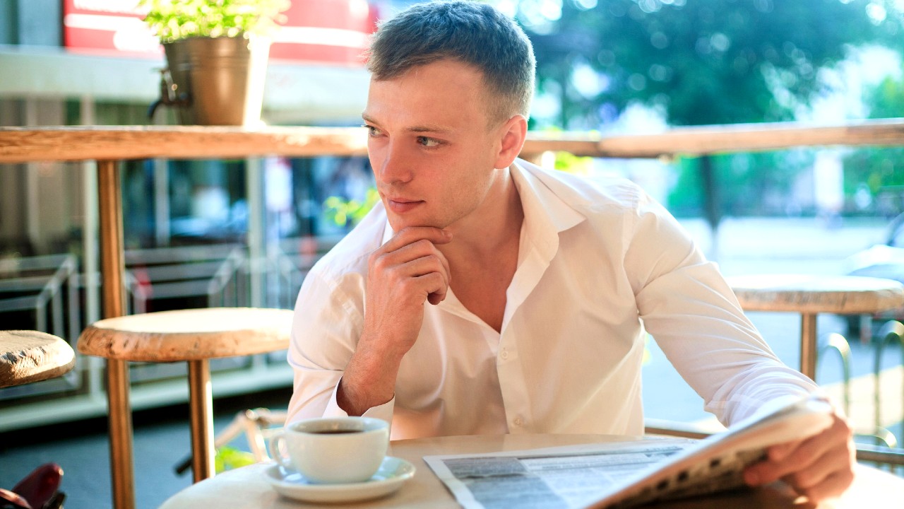 Un uomo in camicia bianca è seduto al tavolo di un bar all'aperto, con un giornale in una mano e il mento appoggiato sull'altra. Una tazza di caffè è sul tavolo di fronte a lui. Guarda pensieroso in lontananza.