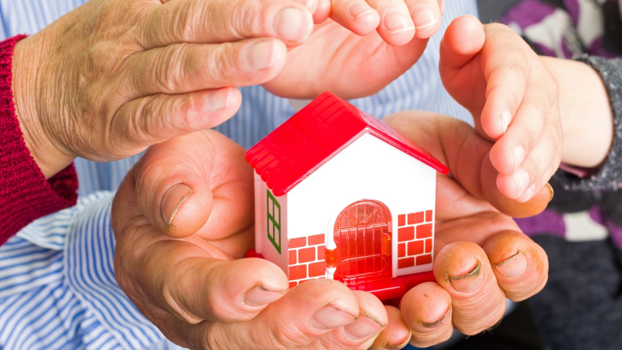 Le mani di un bambino sono incastonate in quelle di un adulto, entrambe cullano una piccola casa giocattolo. La casa bianca con il tetto e la porta rossi simboleggia protezione e cura, proprio come un fondo patrimoniale che salvaguarda i beni di famiglia per le generazioni future.