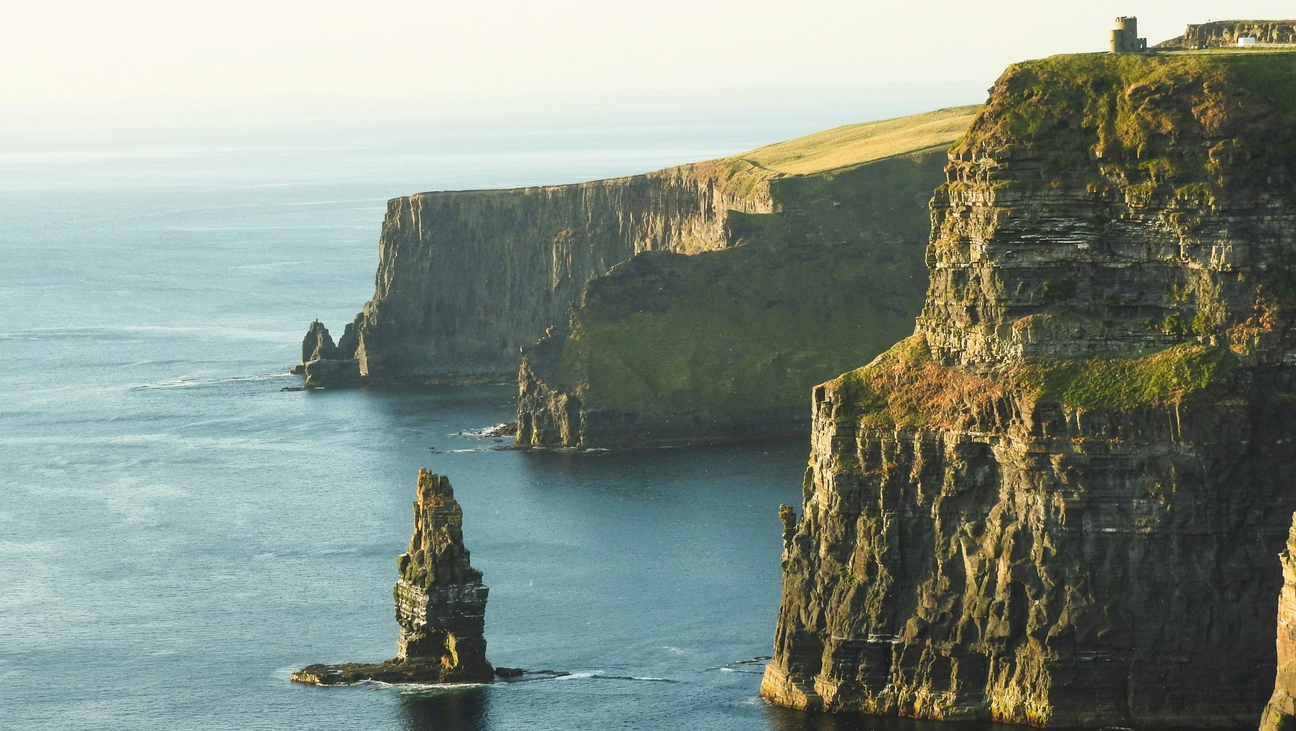 L'immagine mostra le Cliffs of Moher, che mostrano pareti rocciose a strapiombo che si innalzano in modo spettacolare dall'Oceano Atlantico. Le scogliere sono immerse in una luce solare soffusa, con una torre visibile in cima e una vegetazione lussureggiante che ricopre parti del paesaggio roccioso.