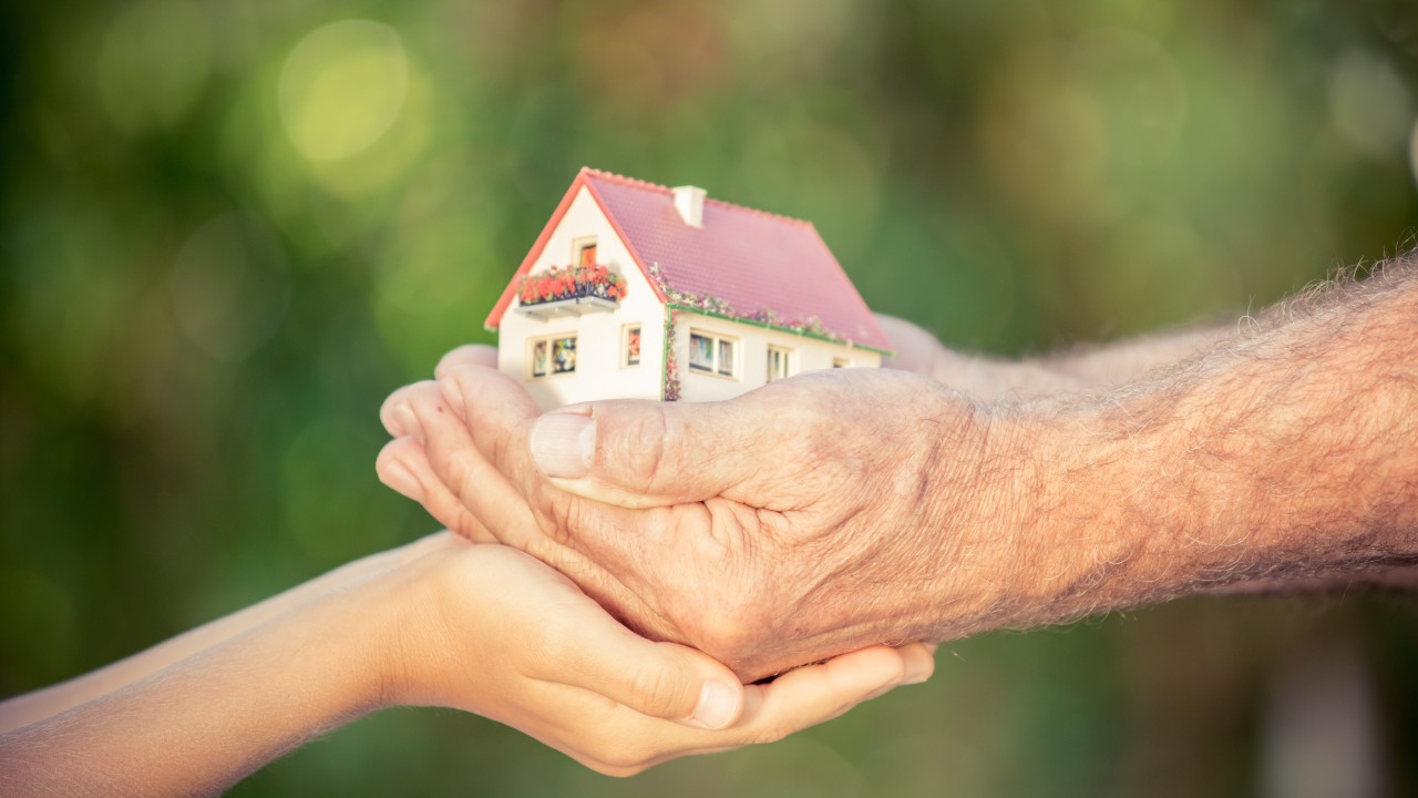 Le mani di un adulto anziano pongono delicatamente un piccolo modello di casa in quelle di una persona più giovane, a simboleggiare il trasferimento della proprietà o dell'eredità. Lo sfondo, delicatamente sfumato con tonalità verdi, suggerisce un ambiente all'aperto in cui la società e i valori condivisi vengono trasmessi tra generazioni.