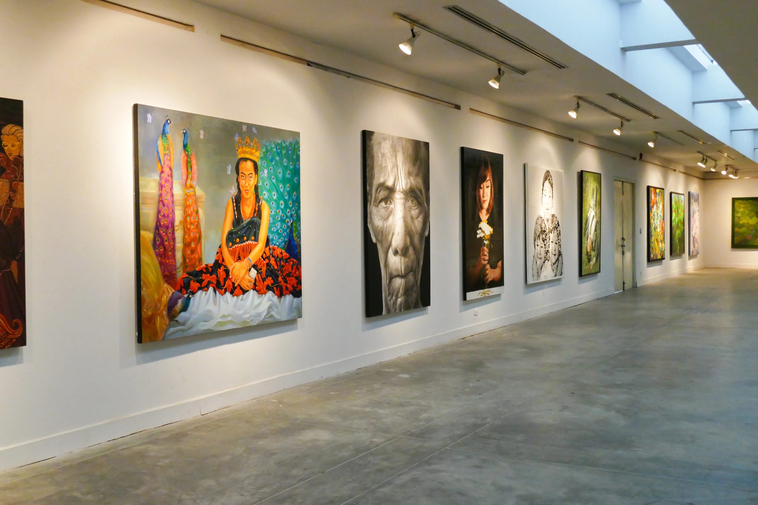 Una galleria d'arte contemporanea, che ricorda un'impresa familiare, presenta un lungo corridoio ben illuminato che espone grandi dipinti colorati. Le opere d'arte includono ritratti e pezzi astratti su pareti bianche. Il pavimento in cemento lucidato completa l'illuminazione a binario e i lucernari soprastanti.