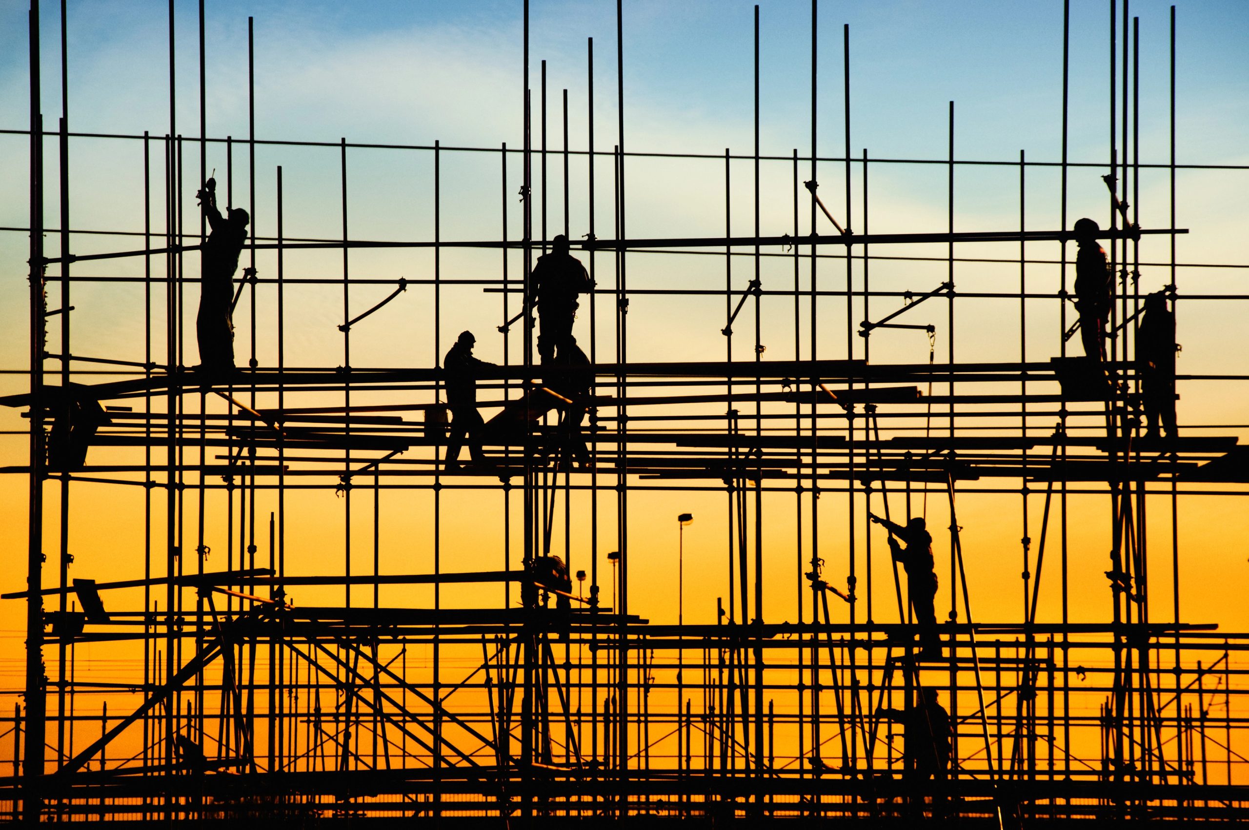 Operai edili in silhouette stanno in piedi e si arrampicano su un'impalcatura in un cantiere durante il tramonto, con un cielo sfumato che passa dal blu all'arancione sullo sfondo.