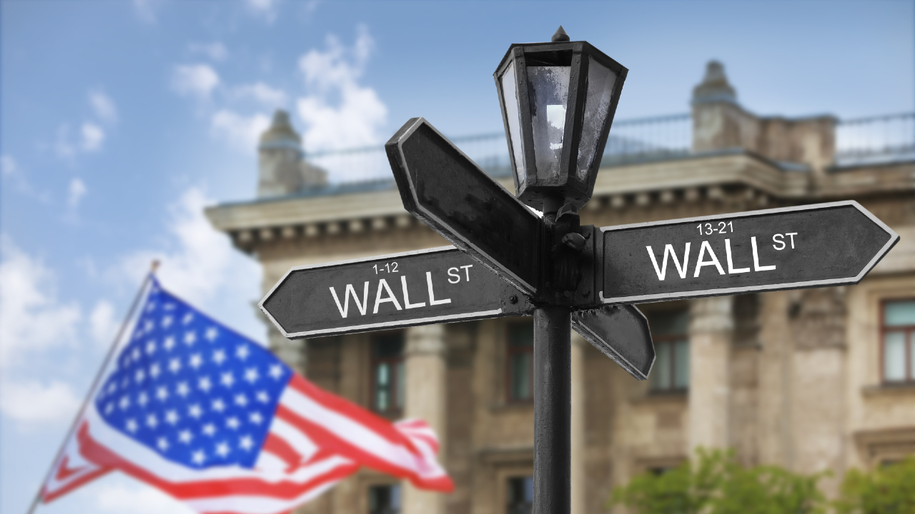 Un cartello stradale nero con la scritta "Wall St" in entrambe le direzioni si erge risoluto davanti a un edificio storico. Sullo sfondo, una bandiera americana sventola dolcemente nella brezza, incarnando Wall Street, il cuore del distretto finanziario di New York City.