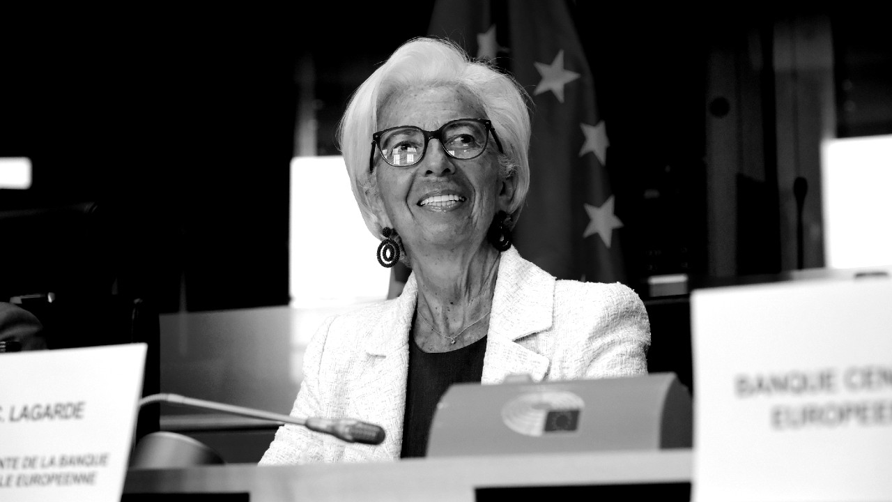Una persona sorridente con i capelli corti e bianchi, occhiali e blazer bianco, siede a un tavolo da conferenza con un microfono. Una bandiera e dei cartellini con i nomi sono esposti lì vicino, a indicare un evento ufficiale.