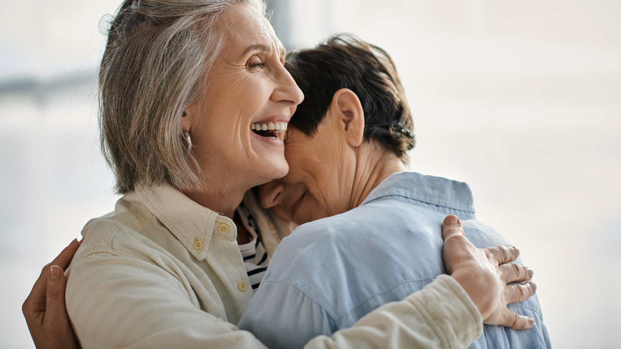 Due donne anziane si abbracciano calorosamente in casa, trasudando felicità e connessione. Con una che sfoggia capelli grigi e l'altra corti riccioli scuri, indossano camicie casual. I loro sorrisi riflettono la gioiosa compagnia che spesso si ritrova negli anni della pensione.