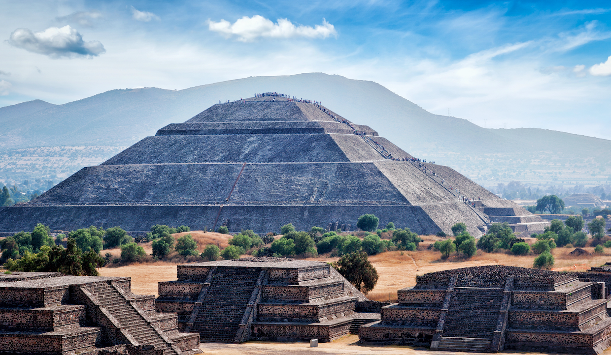 L'immagine mostra la Piramide del Sole a Teotihuacan, in Messico. Una grande struttura in pietra con gradini, circondata da pianure erbose e piramidi più piccole, sotto un cielo azzurro con nuvole sparse e montagne lontane.