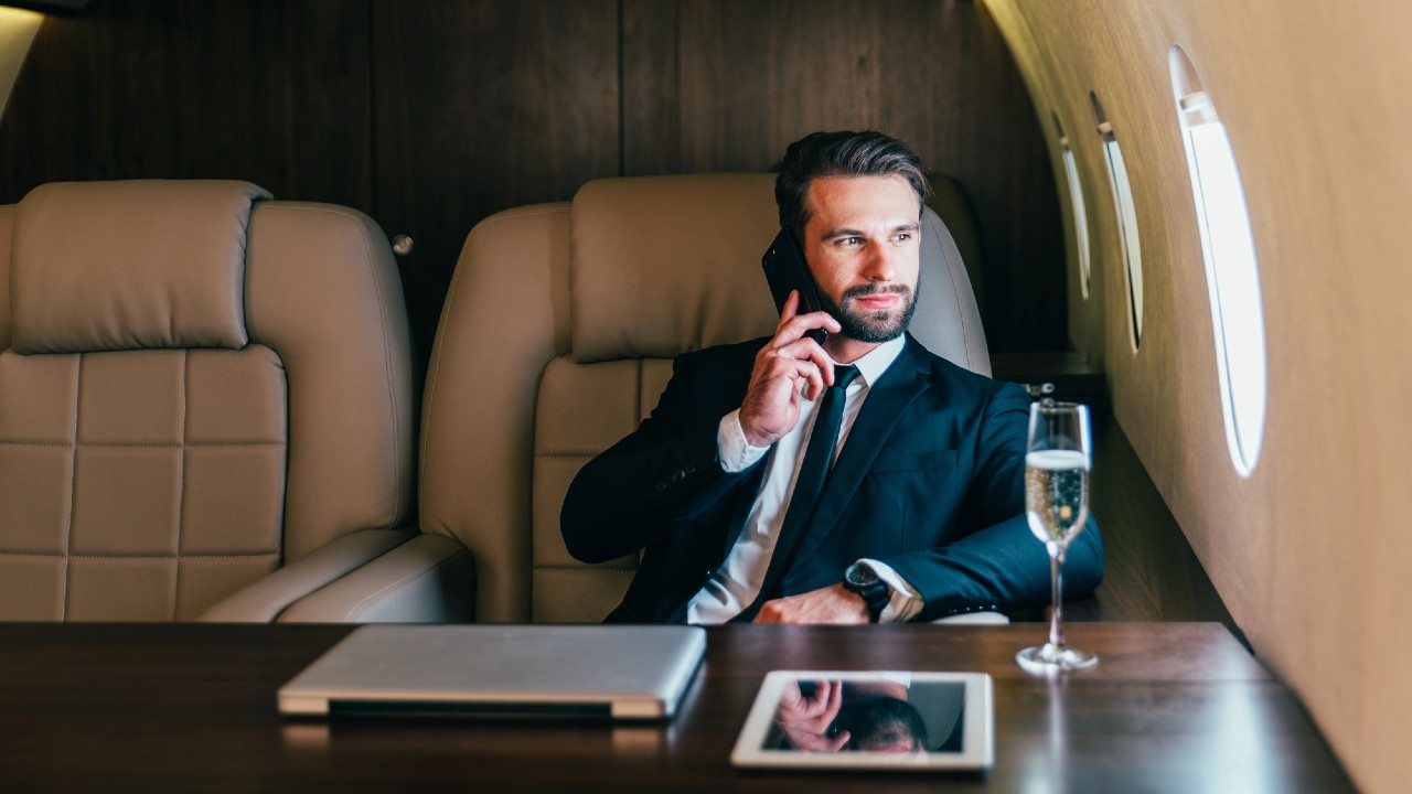 Un uomo in giacca e cravatta parla al telefono mentre è seduto in un jet privato. C'è un portatile sul tavolo di fronte a lui e un bicchiere di champagne. La finestra accanto a lui mostra la luce del giorno.