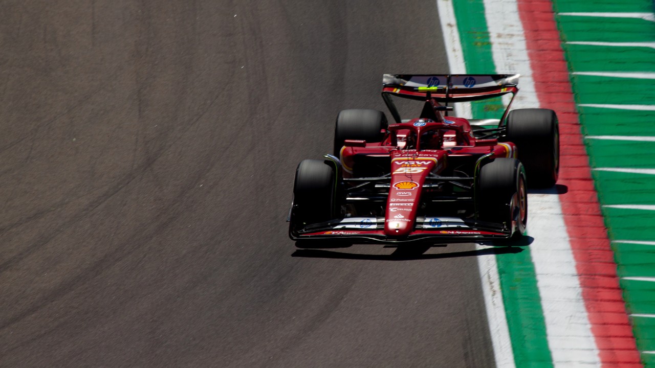 Una monoposto rossa di Formula 1 sfreccia lungo una pista, passando vicino ai cordoli verdi e bianchi. Il pilota, parzialmente visibile, indossa un casco. La vettura ha i loghi degli sponsor ed è immortalata da un'angolazione dall'alto, che ne evidenzia il design elegante.