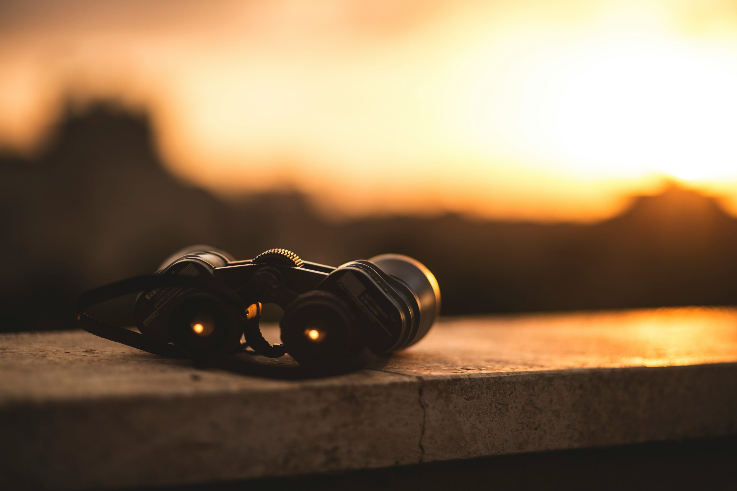 Binocolo appoggiato su una superficie di pietra al tramonto, con uno sfondo sfocato e il caldo chiarore del sole all'orizzonte.