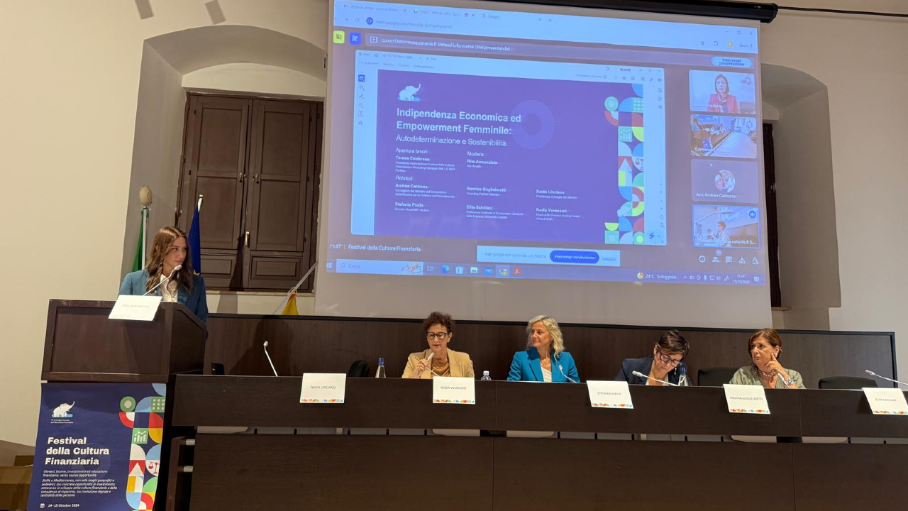 Una donna è in piedi su un podio, e parla al pubblico di "Indipendenza Economica ed Empowerment Femminile". Dietro di lei, una presentazione discute di risparmio gestito. Quattro persone sono sedute a un tavolo sul palco, e ascoltano attentamente.